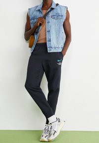 Jeansväst med fransade ärmar, beige undertröja, svarta sweatpants med teal Nike-logga och beige sportskor med grå accenter.