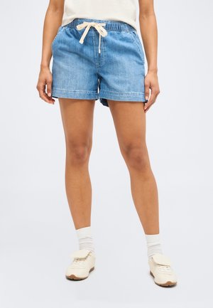 Osoba nosi svetlomodré denimové šortky s bielym sťahovaním, biele ponožky a krémové tenisky, stojí na jednofarebnom pozadí.