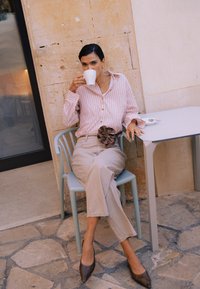 Femme aux cheveux foncés, vêtue d'une chemise à rayures et d'un pantalon beige, assise sur une chaise bleue, buvant dans une tasse blanche dans un café à mur de pierre.