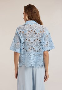 Ljusblå spetsblus med blomstertryck, korta ärmar och en öppen ryggdesign. Slät textur med invecklad utskärningsdetaljering.