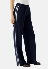 Pantalons larges bleu marine avec des bandes blanches sur les côtés, taille élastique et poches, fabriqués à partir d'un tissu lisse. Convient pour une tenue décontractée.
