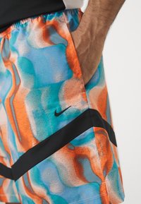 Main d’un homme dans la poche d’un short Nike coloré bleu, orange et turquoise avec une bande en chevron noire et un petit logo Nike noir.
