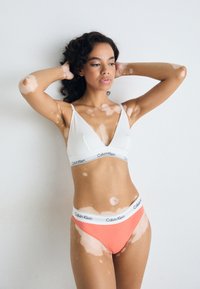 Vrouw met vitiligo in witte en koralen Calvin Klein ondergoed poseert met haar handen achter haar hoofd tegen een effen witte achtergrond.