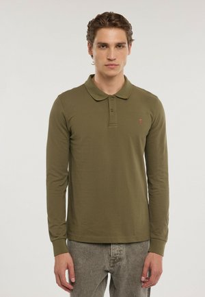 Joven con una camisa polo de manga larga color verde oliva y vaqueros grises, de pie frente a un fondo claro y liso.