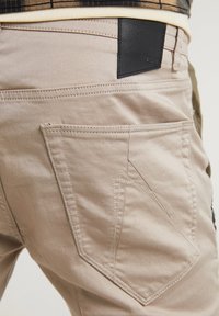 Béžové bavlněné kalhoty s velkou šikmou zadní kapsou s detailním prošíváním a černou koženou nášivkou na pasu.