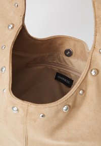 Beige semsket veske med metallnagler langs kanten, har en enkel skulderstropp og en glidelåslomme inni. Merkelappen er synlig.