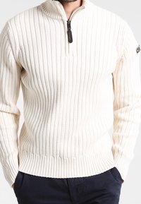 Ljuscremefärgad tröja med horisontell ribbning, hög krage och fyrdelaresdragkedja. Har ett märke på vänster ärm för extra detalj.