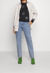 Gestreepte witte en zwarte jas over een zwarte trui met bloemenborduursels, gecombineerd met lichtblauwe jeans en groene blokhak enkellaarsjes.