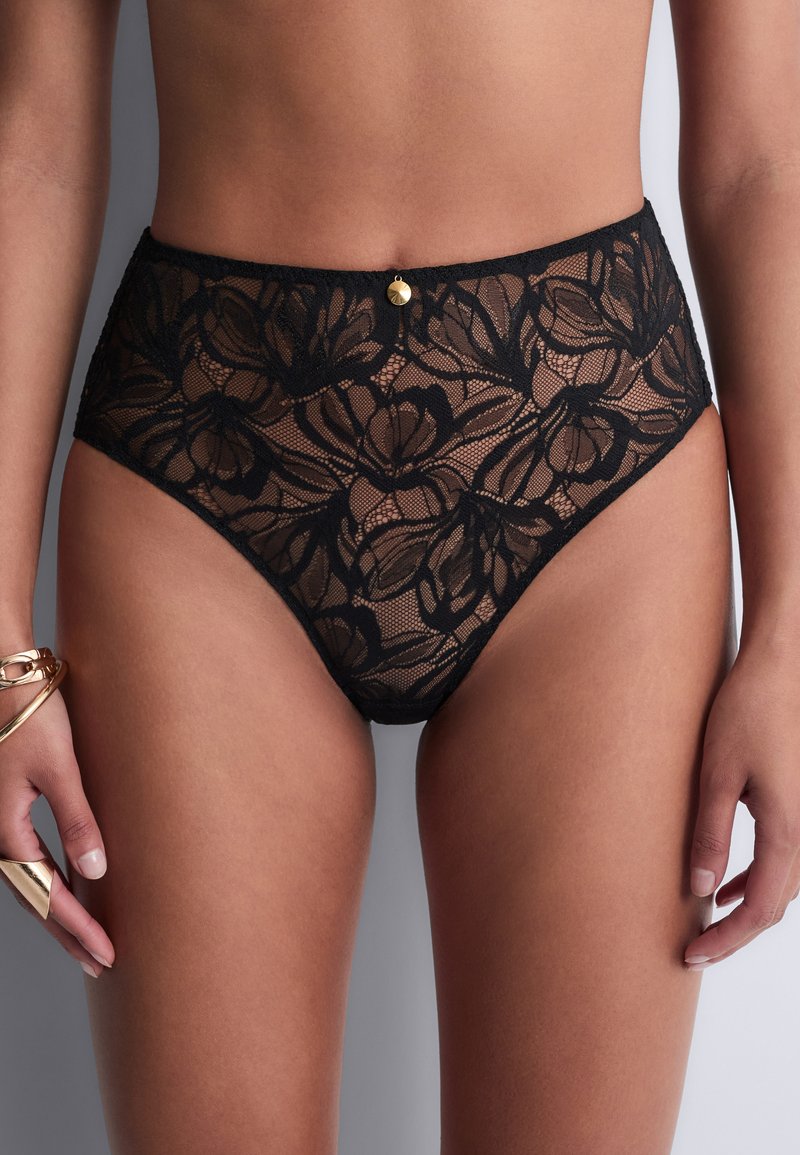 Sous-vêtements en dentelle noire taille haute avec motif floral et petit pendentif doré, portés par une personne avec des bracelets et une bague à la main gauche.