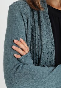 Närbild av en person som bär en strukturerad blå stickad cardigan över en svart topp, med vänster arm korsad och hand resting på överarmen.