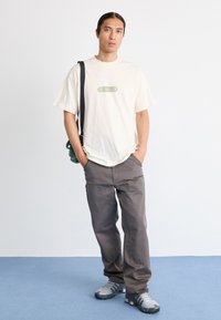 Joven con cabello largo viste una camiseta gráfica blanca, pantalones cargo grises, zapatillas grises y lleva una pequeña bolsa cruzada verde, de pie sobre un suelo azul.