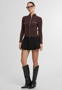 Brązowy prążkowany sweter zapinany na zamek z wysokim kołnierzem, w połączeniu z czarną plisowaną spódnicą i czarnymi skórzanymi botkami za kolano. Widoczny jest wyraźny detal z logo.