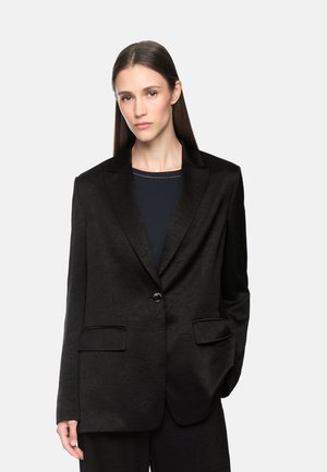 Woman with long dark hair wearing a black textured blazer over a dark top, standing with one hand in pocket against plain background.