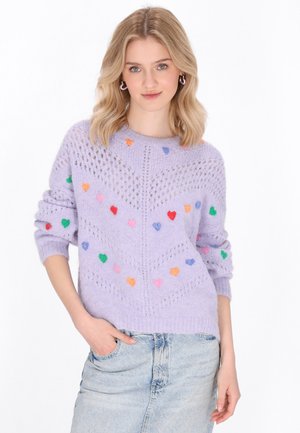 Lila gestrickter Pullover mit einem Muster aus bunten Herzformen, offenen Maschen und lässigen Ärmeln. Kombiniert mit einem Jeansrock.