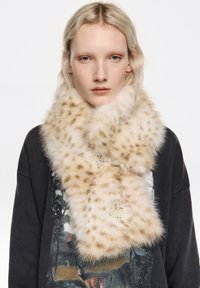 Bufanda de piel sintética con un estampado beige y marrón moteado, con una textura suave y botones redondos. Se lleva sobre una sudadera gráfica oscura.