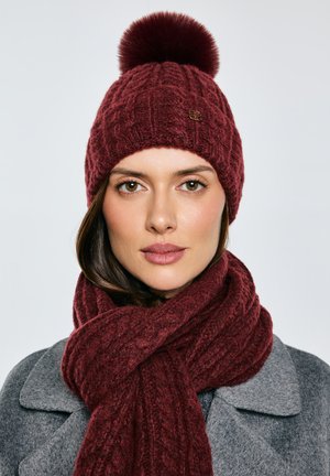 Žena ve vínově červené pletené čepici s bambulí a odpovídajícím šálou, šedém kabátu, čelem vpřed s neutrálním výrazem na světlém pozadí.