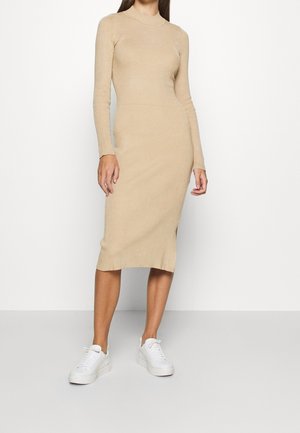Vrouw draagt een beige, geribbelde midi-jurk met lange mouwen, gecombineerd met witte sneakers, staand tegen een effen lichte achtergrond.