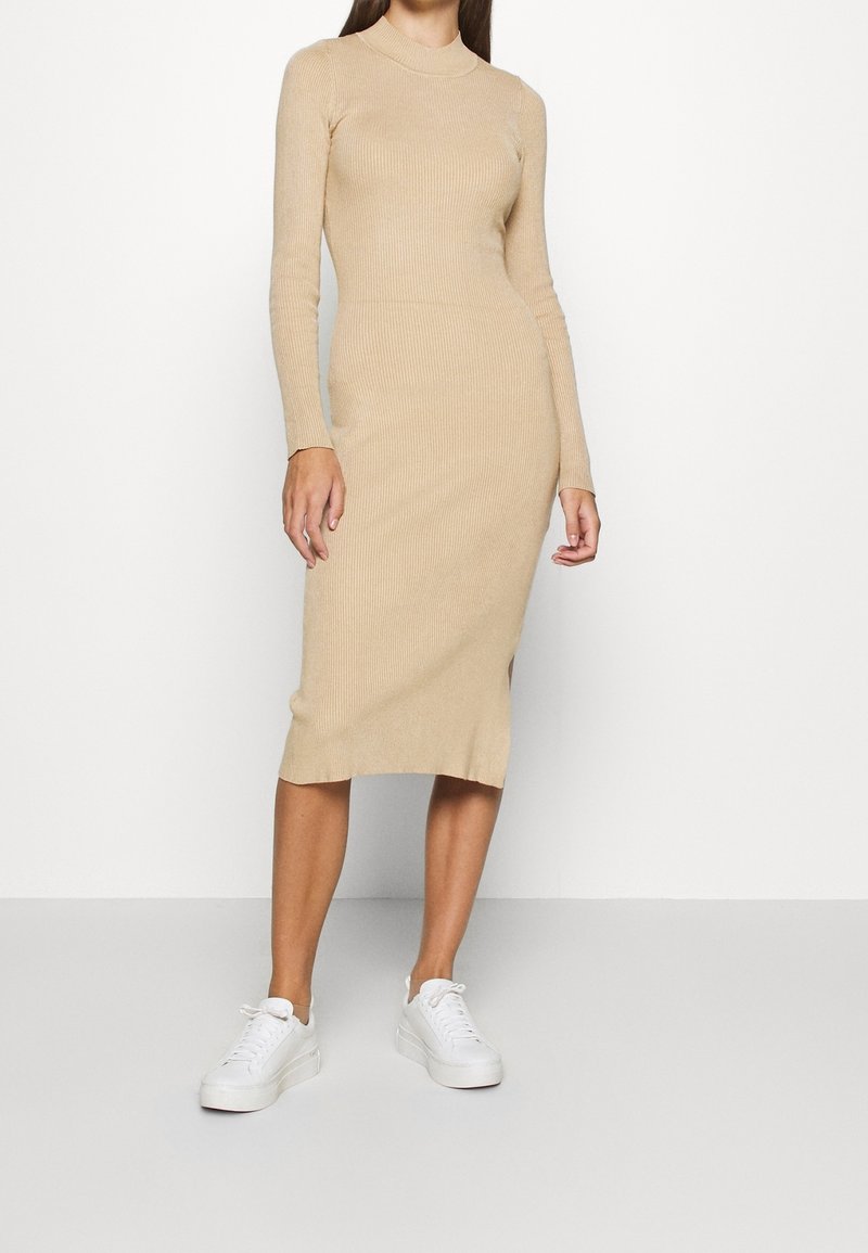 Femme portant une robe midi beige à manches longues et côtelée, associée à des baskets blanches, debout devant un fond clair uni.