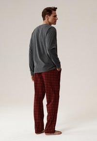 Szara koszula z długim rękawem, w połączeniu z czerwono-czarnymi, w kratkę spodniami piżamowymi, o luźnym kroju i bocznymi kieszeniami. Na boso, widok z tyłu.