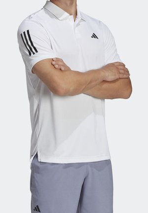 Hombre vestido con polo blanco de manga corta con logo negro y rayas en la manga, y pantalones cortos gris claro, con los brazos cruzados sobre el pecho.