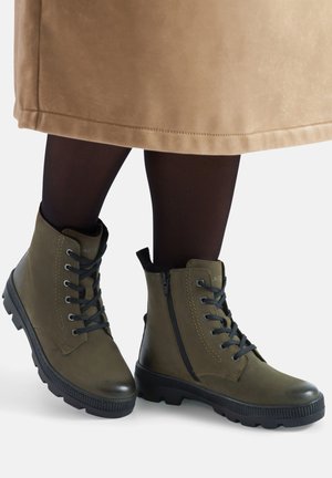 Oliwkowozielone skórzane botki za kostkę z czarnymi sznurowadłami, bocznym zamkiem i teksturowaną gumową podeszwą, noszone z ciemnymi rajstopami i beżową spódnicą.