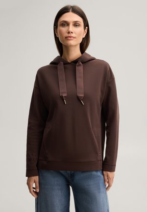 Frau trägt einen braunen Hoodie mit breiten Kordeln und blauen Jeans, schaut nach vorne vor einem schlichten hellen Hintergrund.
