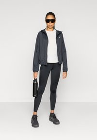 Μαύρο ελαφρύ μπουφάν με κουκούλα πάνω από λευκό πουκάμισο, συνδυασμένο με μαύρα κολάν και μαύρα αθλητικά παπούτσια. Κρατάει μια μαύρη παγούρι νερού.