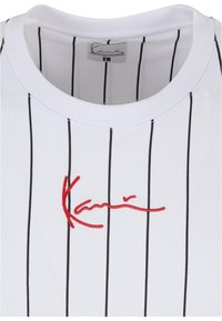 Hvid t-shirt med rund hals og tynde lodrette sorte striber samt rød broderet signatur henover brystet.