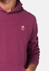 Fioletowa bluza z kapturem wykonana z miękkiego materiału, z małym wyszywanym logo z koniem i napisem "POLO SYLT" na lewej piersi.