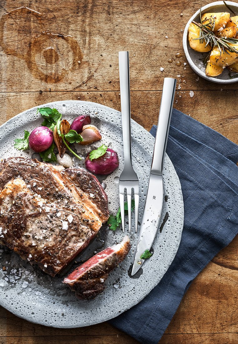 Graue gesprenkelte Platte mit einem Stück gebratenem Fleisch, garniert mit Kräutern und Gemüse. Edelstahlmesser und -gabel auf einer blauen Stoffserviette.