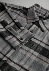 Szara flanelowa koszula w kratę z czarnymi i brązowymi akcentami, z guzikami na mankietach i kieszenią na piersi. Miękka struktura i kołnierzyk.