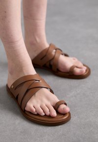 Brown leather slide sandals with crossed straps and a toe loop. Flat sole, minimal cushioning, and smooth texture, shown on bare feet.