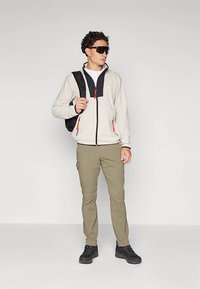 Muškarac s naočalama za sunce, svijetlo bež jakna s narančastim patentnim zatvaračima, kaki hlače, crne cipele i crni ruksak, stoji nasuprot jednobojnom bijelom zidu.