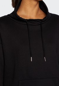 Sudadera negra con cuello alto, cordones ajustables con puntas metálicas y un bolsillo delantero. Textura de tela suave, diseño casual.