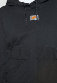 Svart hoodie med två stora framfickor, texturerad accenttyg och en liten, färgglad Nike-logotag på bröstet. Har dragsko.