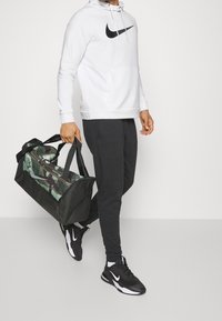 Biała bluza z kapturem z czarnym logo Nike, czarne dresy i czarne sneakersy Nike. Niesie czarną torbę podróżną w kamuflażu w odcieniach zieleni z podwójnymi uchwytami.