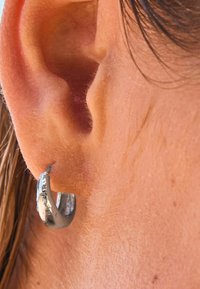 Silberner Creolenohrring mit strukturiertem Finish, der eine kreisförmige Form und eine schmale Öffnung aufweist und im Ohrläppchen liegt.