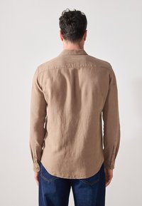 Camicia in lino marrone con polsini abbottonati, colletto all'inglese e orlo curvo, indossata sopra jeans in denim blu, vista da dietro.