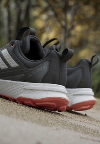 Chaussures de trail Adidas Terrex gris foncé avec semelles intermédiaires blanches et semelles extérieures rouges sur une surface de gravier, fond naturel flou.