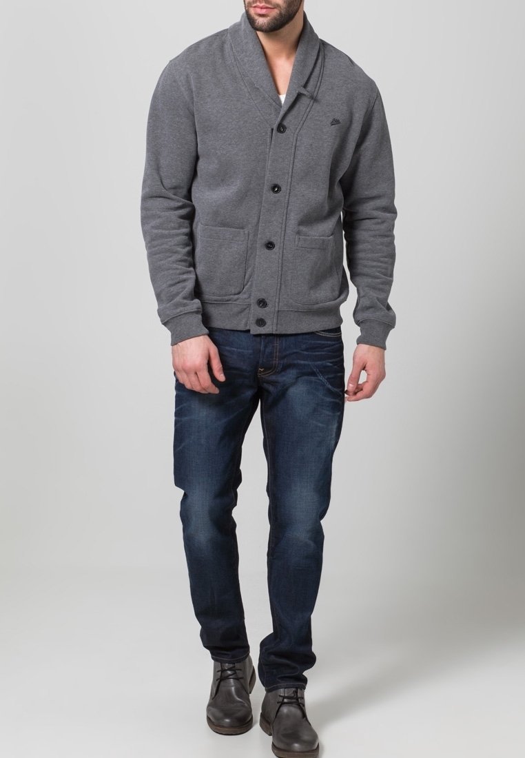 Grå cardigan med sjalkrage, knappstängning, två framfickor, bärs över blå jeans och bruna stövlar. Slät yta, stickat material.