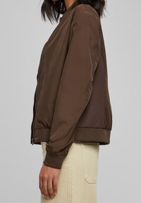Chaqueta bomber marrón con cremallera frontal, puños acanalados y panel texturizado en la espalda. Material suave con un acabado ligeramente brillante.