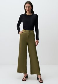 Zwart geribd langwerpig topje gecombineerd met olivegroene high-waisted wijde broek. Model draagt zwarte strapsandalen. Zachte stoftextuur.