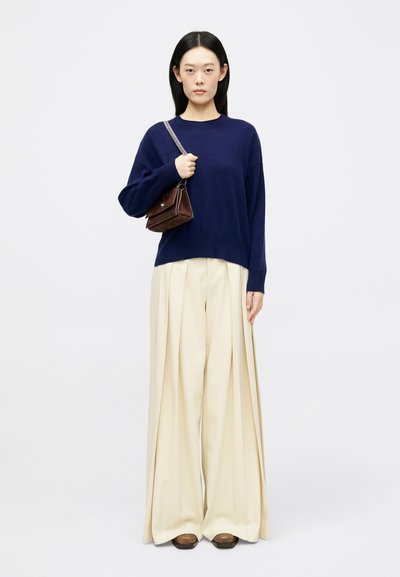 Donna con capelli neri lisci, che indossa un maglione blu navy, pantaloni larghi color crema, scarpe marroni e tiene una piccola borsa marrone con una tracolla a catena.