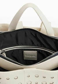 Bolso blanco con diseño perforado en el exterior, forro de tela negra y bolsillo con cremallera que incluye una etiqueta con el logo en el interior.
