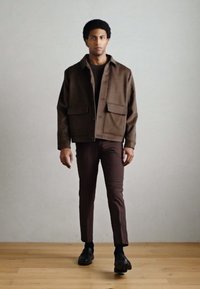 Casaco castanho em mistura de lã com dois bolsos frontais, usado sobre uma camisola escura. Calças castanhas de corte justo e sapatos pretos. Fundo neutro.