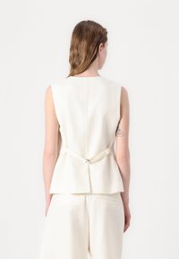 Krämfärgad ärmlös topp med peplumdesign och bakband. Tillverkad av ett mjukt material med minimala sömnadsdetaljer.