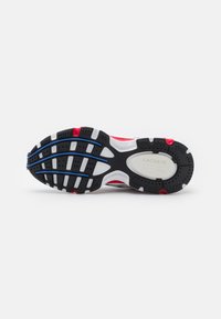 A Lacoste sportcipő talpa fekete gumiból készült, amely több irányban is tapadó mintázattal rendelkezik, piros és kék díszítésekkel, valamint egy fehér logó részlettel.