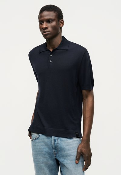 Άνδρας που φορά μαύρο πόλο με κοντά μανίκια και δύο κουμπιά και ανοιχτό μπλε τζιν, στέκεται μπροστά σε απλό ανοιχτό φόντο.