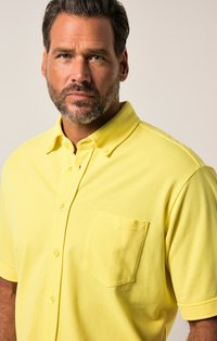 Camisa polo amarilla de mezcla de algodón, con mangas cortas, cuello abotonado y un bolsillo en el pecho. Textura suave, con detalles en botones.