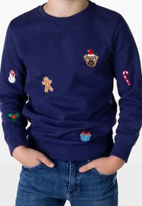 Dziecko w granatowym swetrze z motywami świątecznymi, w tym buldogiem francuskim w czapce Mikołaja, piernikiem, bałwankiem, cukrową laską, ostrokrzewem i pudełkiem prezentowym.